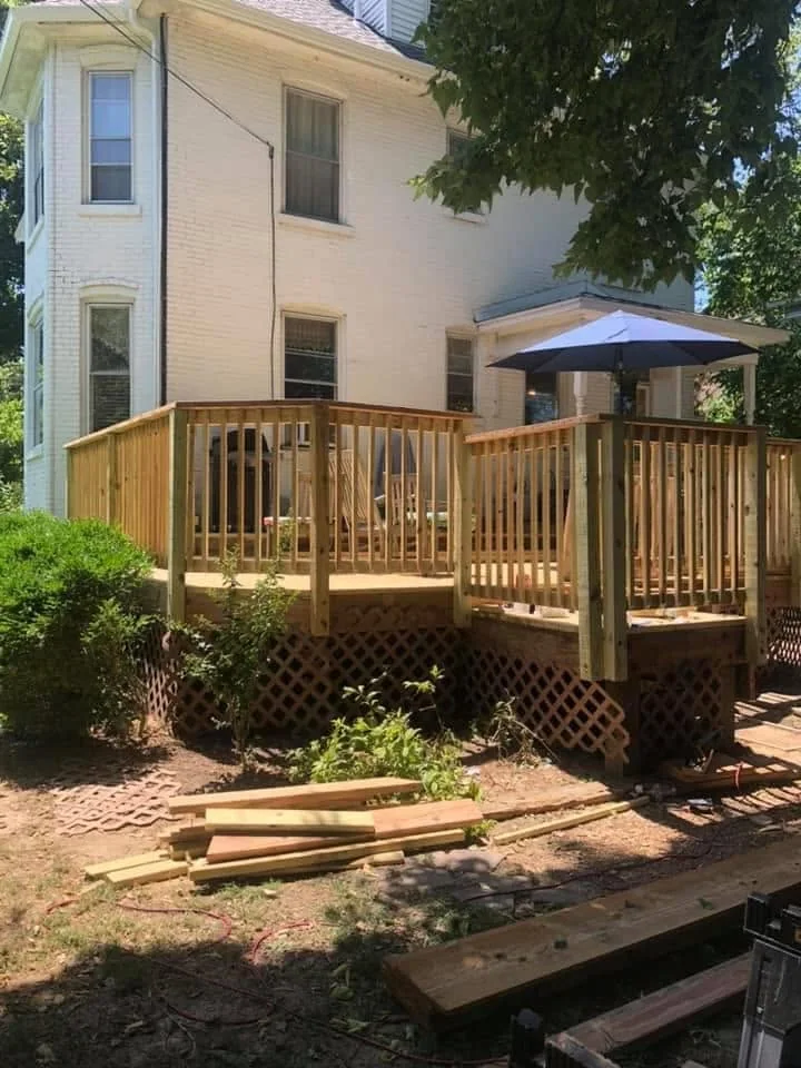 Raised wood deck with railings and lattice skirting beside a white brick home, shaded by a patio umbrella